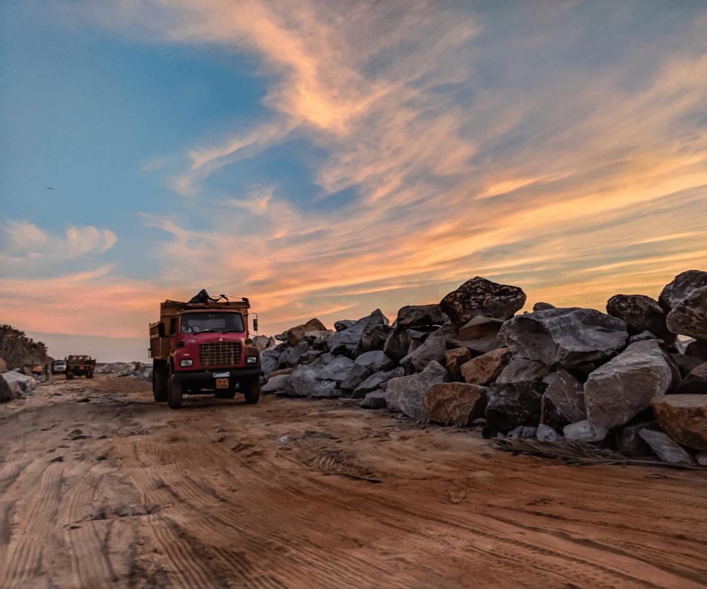 A red dump truck moves past rocks against an evening sky