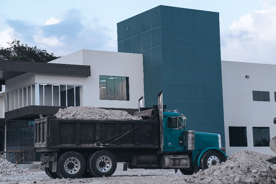 A dark teal and black dump truck holds rubble at a construction site with a building in the background