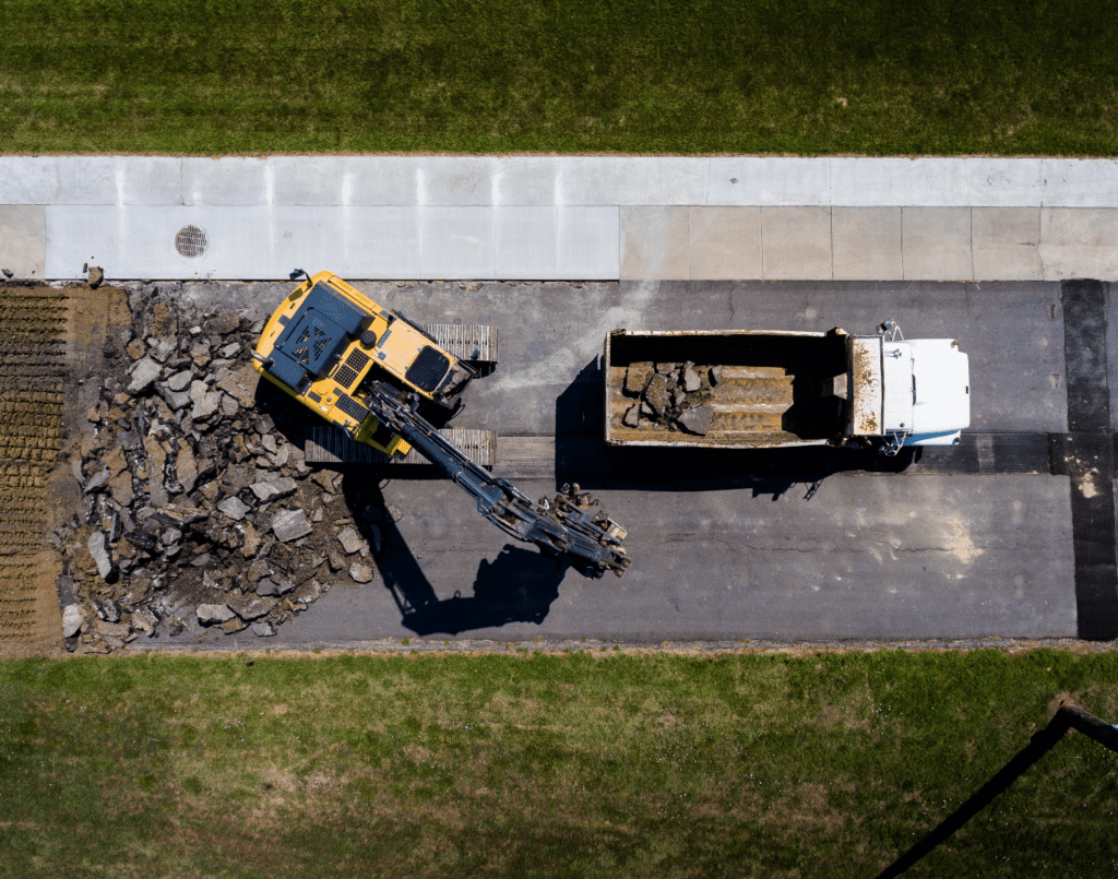 Aerial photography of a yellow excavator breaking the road beside a white dump truck by a patch of green grass