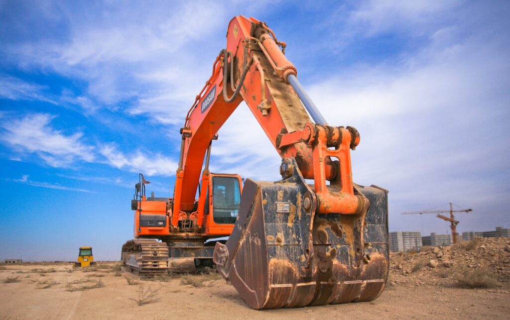 A large orange excavator with a long arm and bucket viewed from a low angle, with white clouds in a blue sky above.