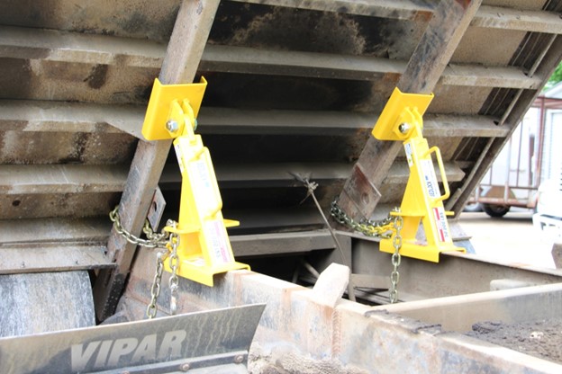 A pair of yellow truck bed locks, as mentioned in this BedLock Safety Products guide, holding up the raised dump bed of a dump truck