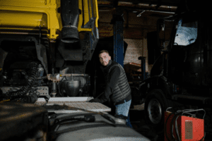 A mechanic standing beside a raised dump truck bed inside a workshop.