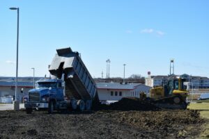 Dump truck unloading dirt in front of an industrial building, showing active material handling during heavy-duty truck operations.