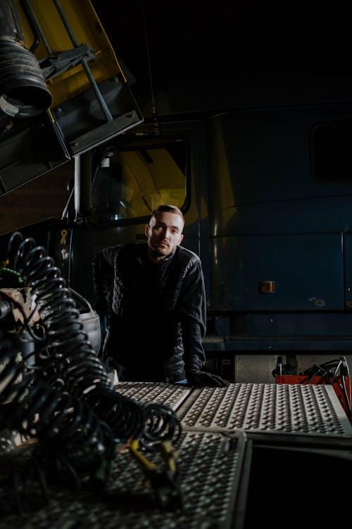 A mechanic standing beside a raised dump truck in a repair shop, checking the vehicle during maintenance.