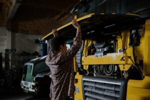 Technician inspecting a truck engine with the hood open inside a repair shop, following safety protocols.