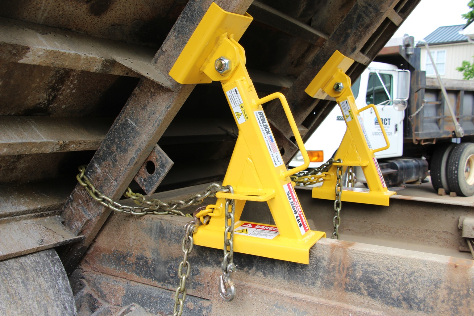 Close-up of two yellow BedLock safety devices installed with chains beneath a raised dump truck bed
