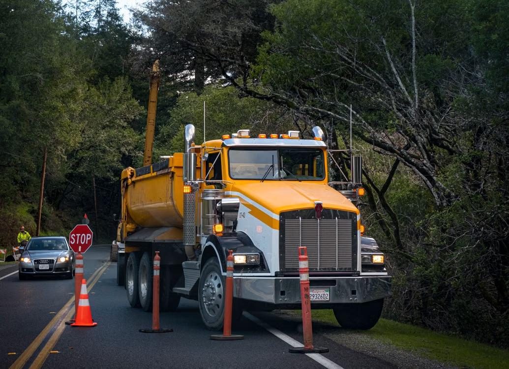 Yellow dump truck driving on a paved road past a stop sign and traffic cones