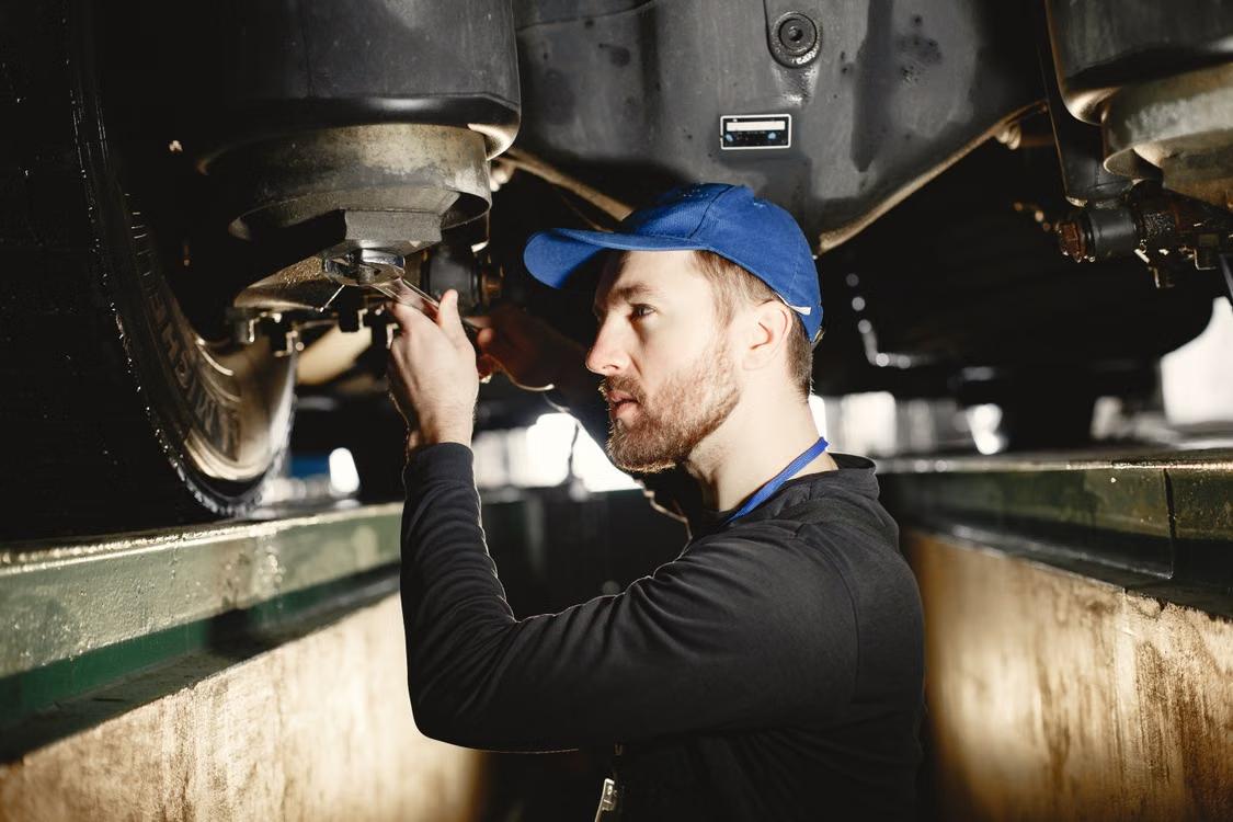 Technician in a blue cap inspecting suspension components underneath a truck
