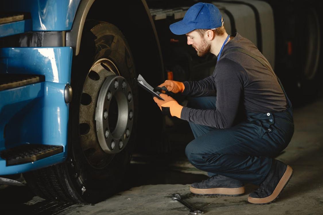 Technician kneeling to inspect a truck wheel while holding a clipboard and pen